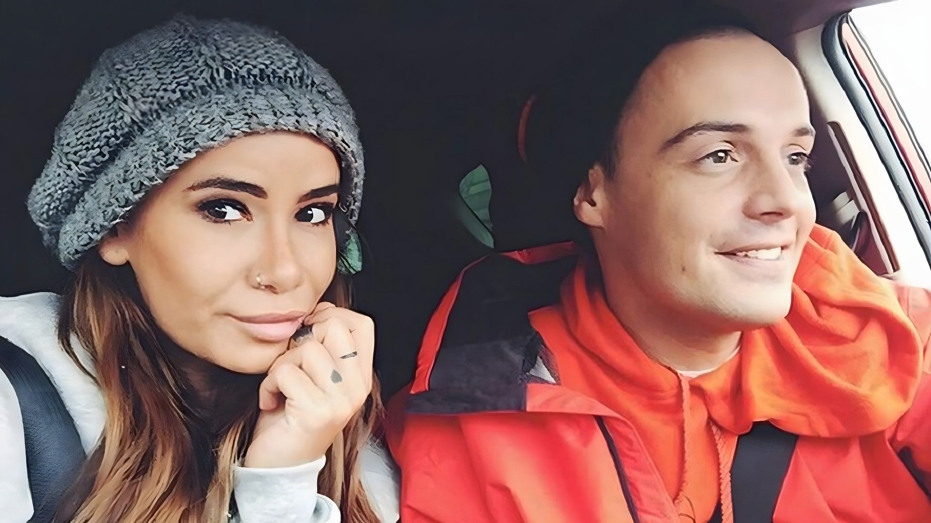 Девушка в серой кофте и вязаной шапке и молодой человек в краснной куртке едут в автомобиле, селфи