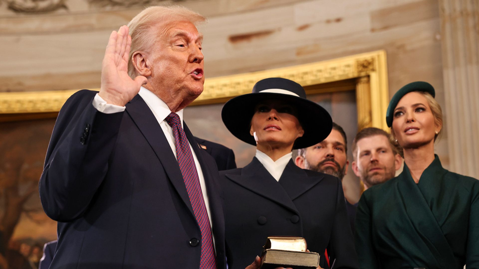Дональд Трамп принимает присягу на трибуне, сзади него стоит Мелания Трамп в шляпе с очень широкими полями
