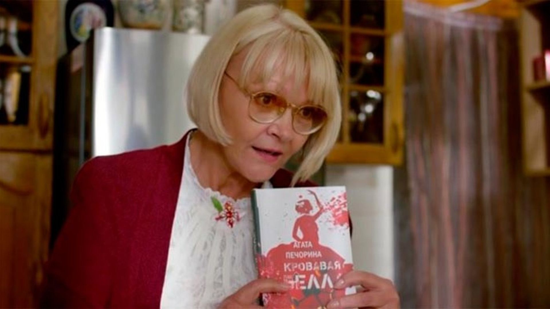 Марина Дюжева с прической каре, в красной кофте и белой блузке, держит в руках книгу