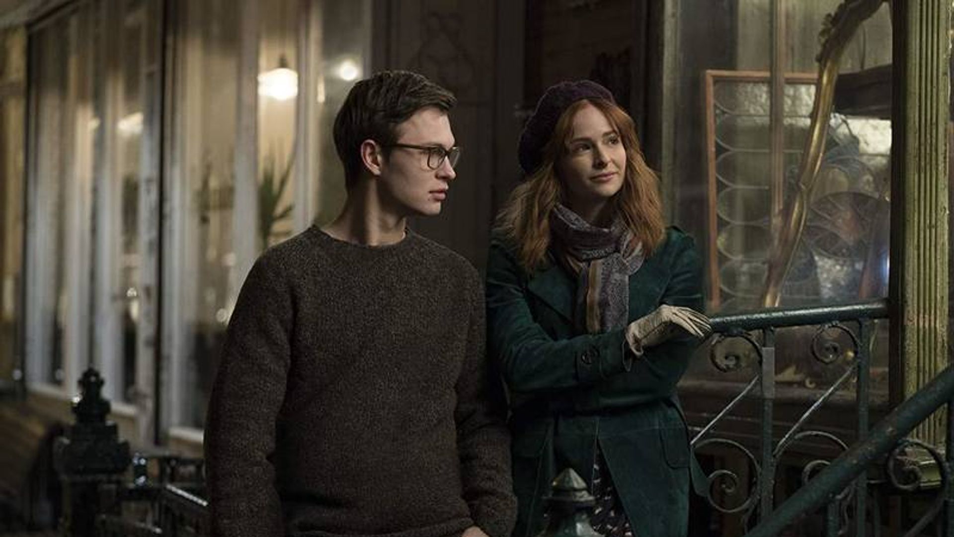 Молодой мужчина в очках и женщина в пальто и берете разговаривают, стоя у перил на улице вечером