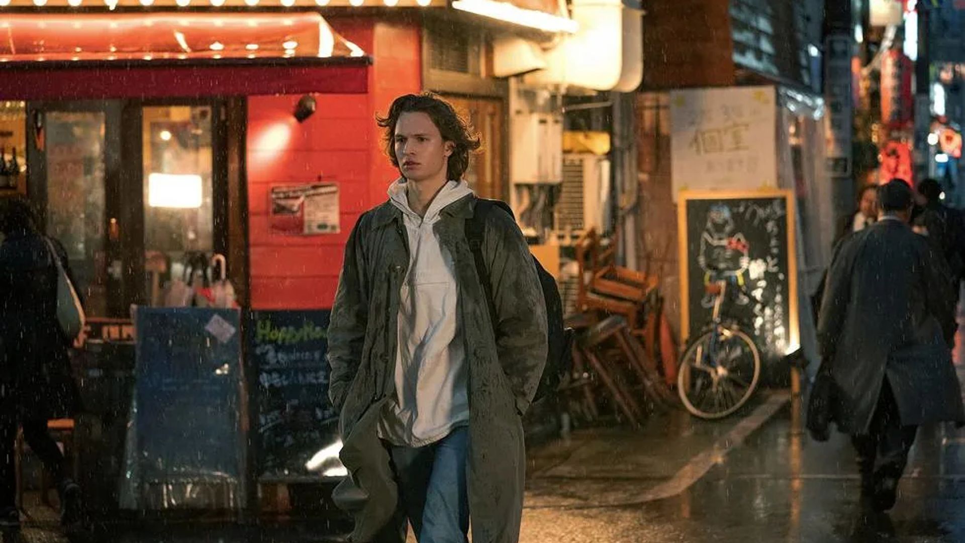 Энсел Эльгорт в плаще и худи идёт по дождливой улице мимо ярко освещённых витрин и ресторанов