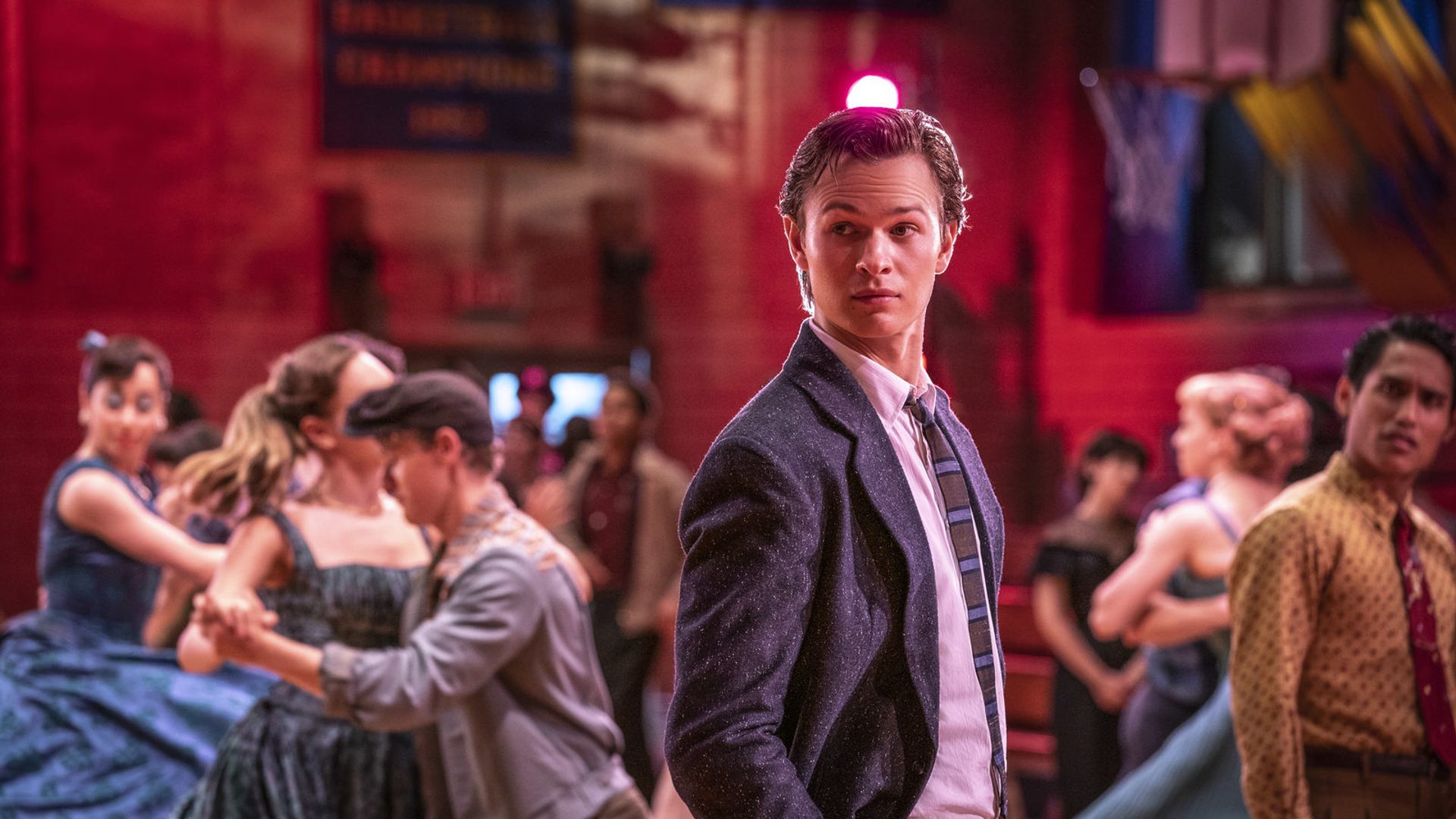 Энсел Эльгорт в костюме и галстуке стоит на танцевальном вечере, вокруг пары кружатся в танце