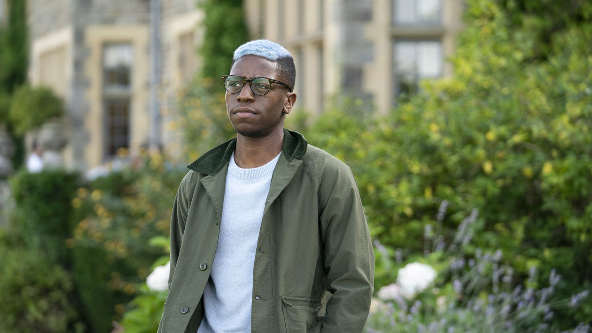 Дэвид Джонссон в очках и с голубыми волосами стоит на улице, одетый в зеленую куртку