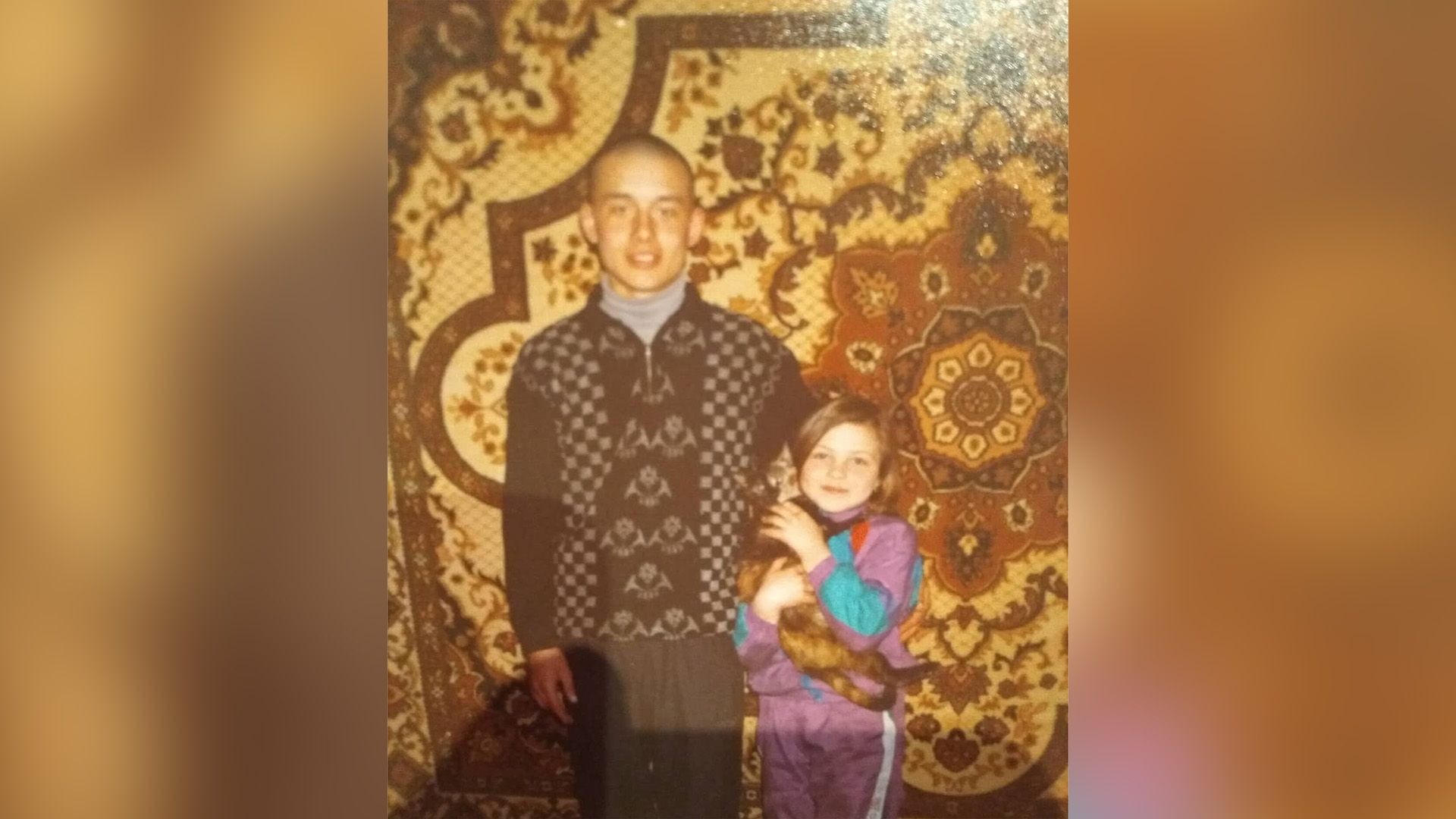 Анастасия Тюнина с кошкой на руках стоит рядом со старшим братом на фоне ковра