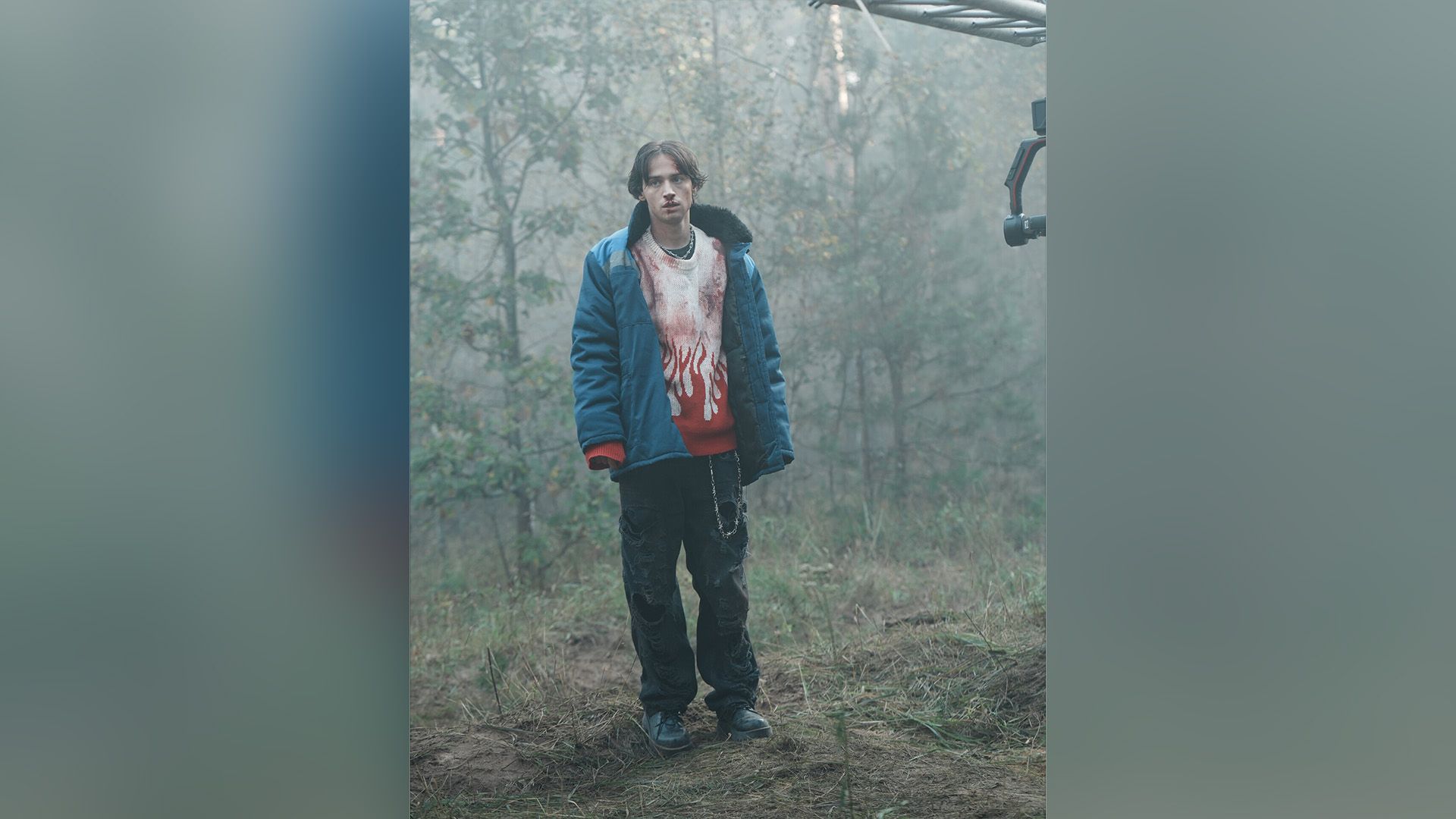 Андрей Мартынов в рваных джинсах и синей куртке стоит в лесу, на его лице и свитере кровь
