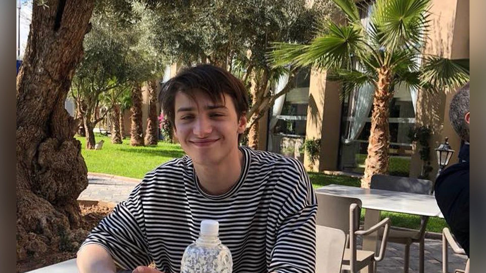 Улыбающийся Андрей Мартынов сидит за столом на улице на фоне пальм