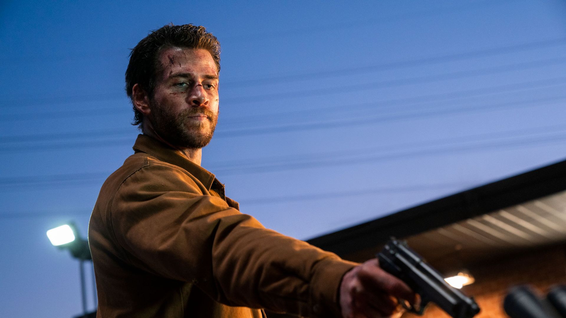 Лиам Хемсворт в коричневой куртке держит пистолет