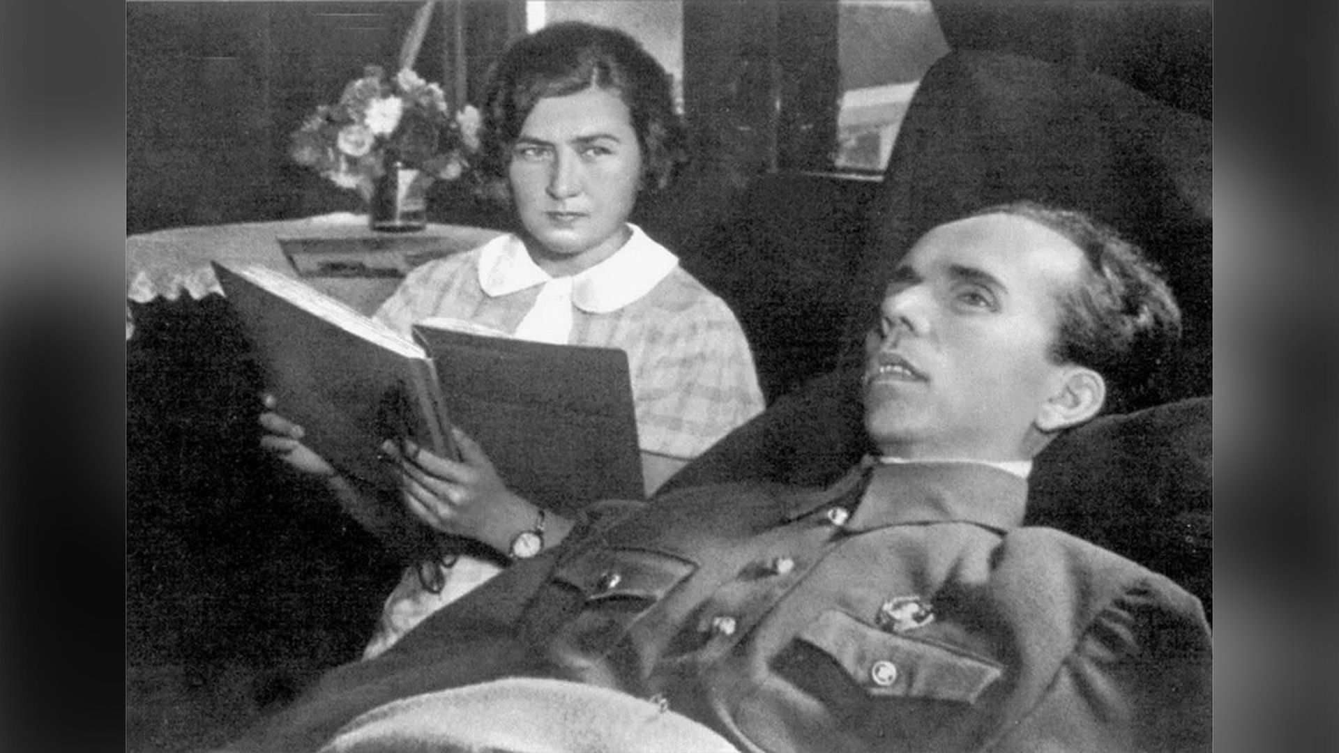 Николай Островский лежит на кровати, рядом сидит его жена Раиса с открытой книгой в руках