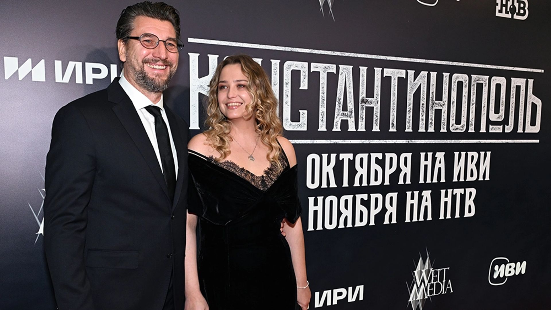 Устюгов и Шиловская в вечерних нарядах держатся за руки на фоне стены с надписью Константинополь