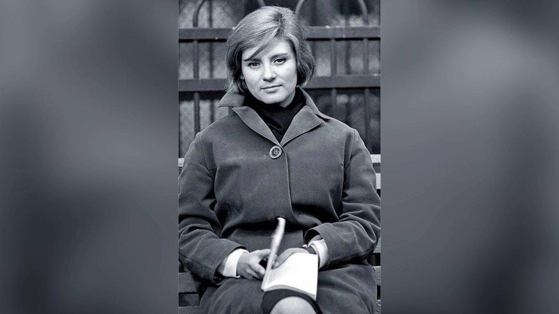 Актриса Евгения Уралова в пальто сидит на скамейке, держа в руках книгу