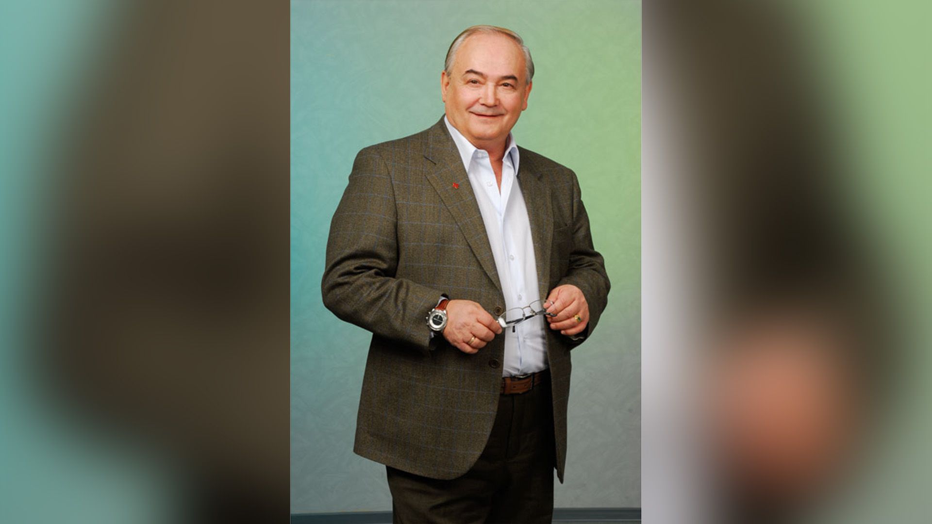 Всеволод Шиловский в коричневом пиджаке стоит, держа в руках очки