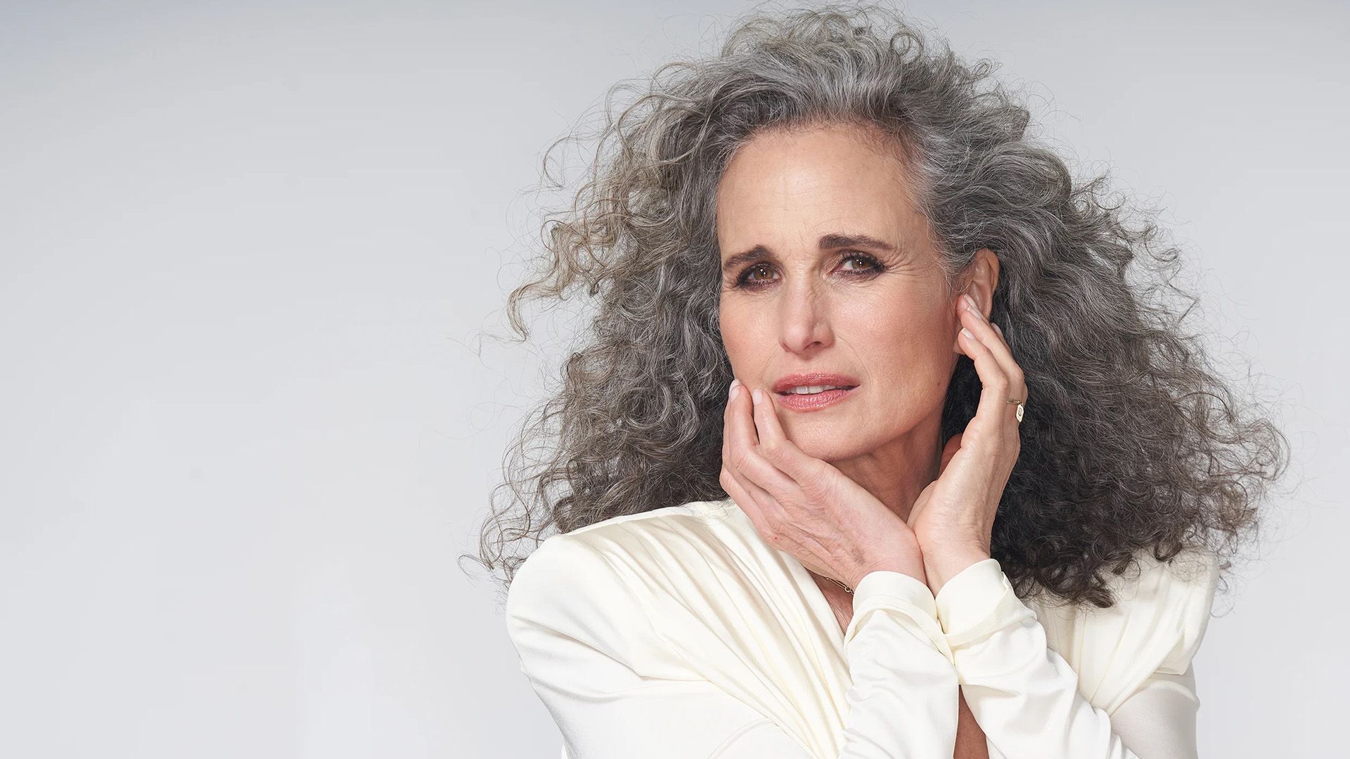 Актриса Энди МакДауэлл с развевающимися седыми кудрями, одетая в белый пиджак, касается руками своего лица