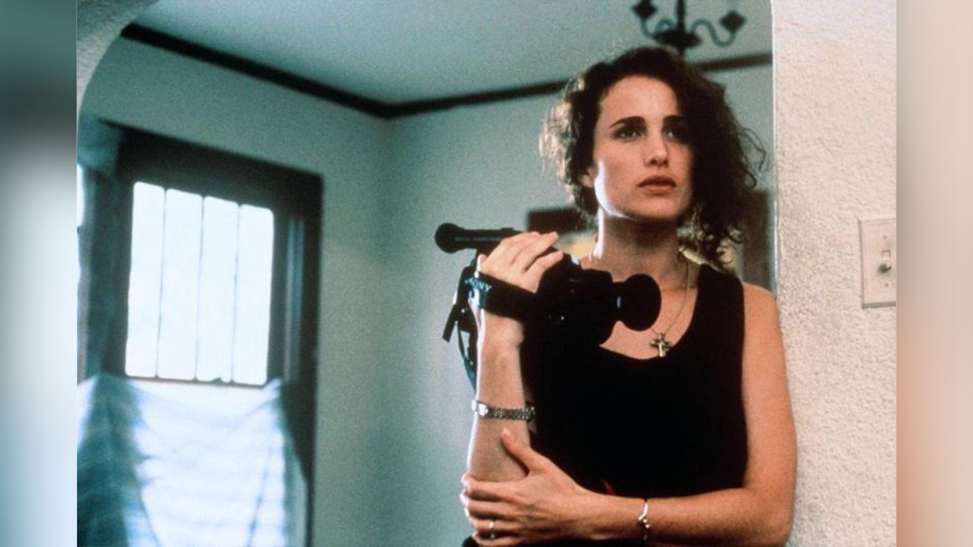 Энди МакДауэлл в чёрной майке стоит в дверном проёме, держа в руках видеокамеру