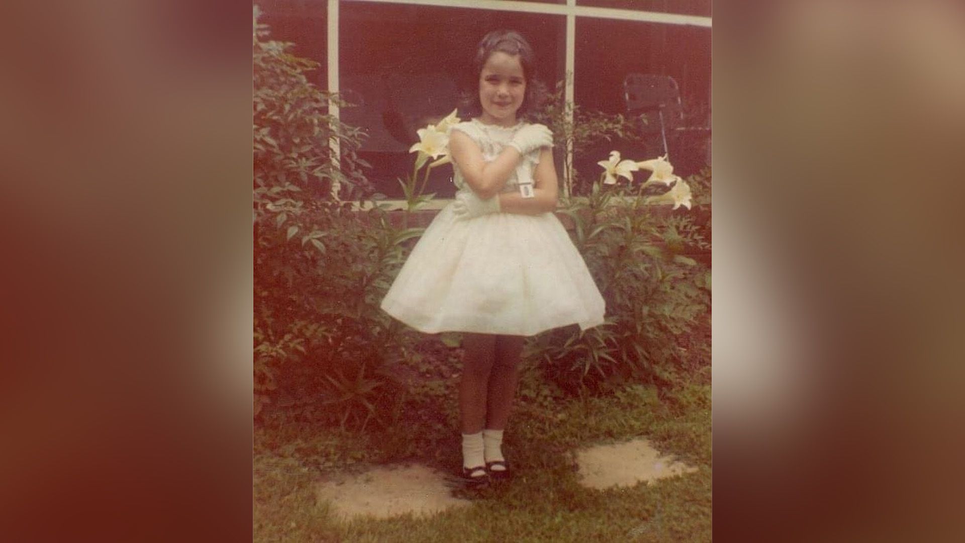 Маленькая Энди МакДауэлл в белом платье с пышной юбкой и белых перчатках стоит в саду с белыми лилиями