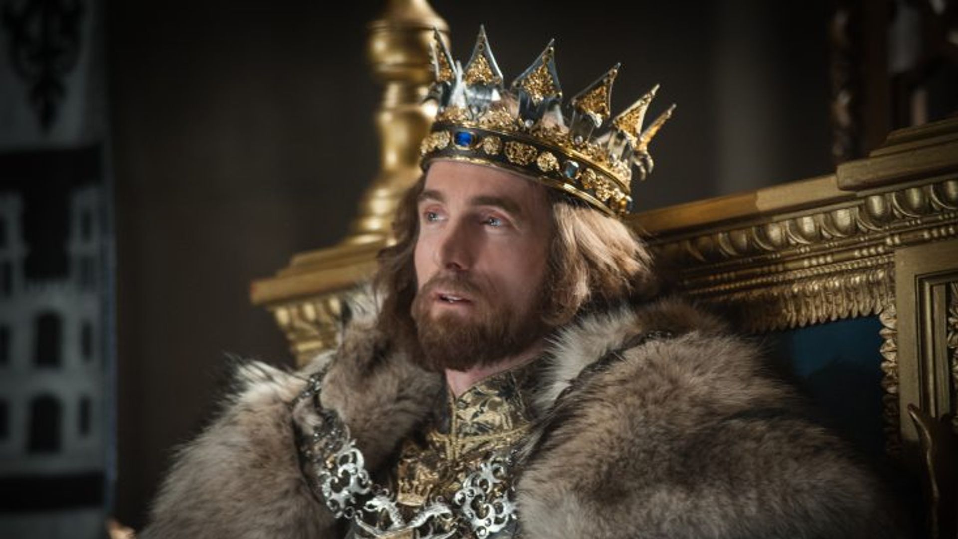 Шарлто Копли в костюме короля сидит на троне с отстранённым выражением лица