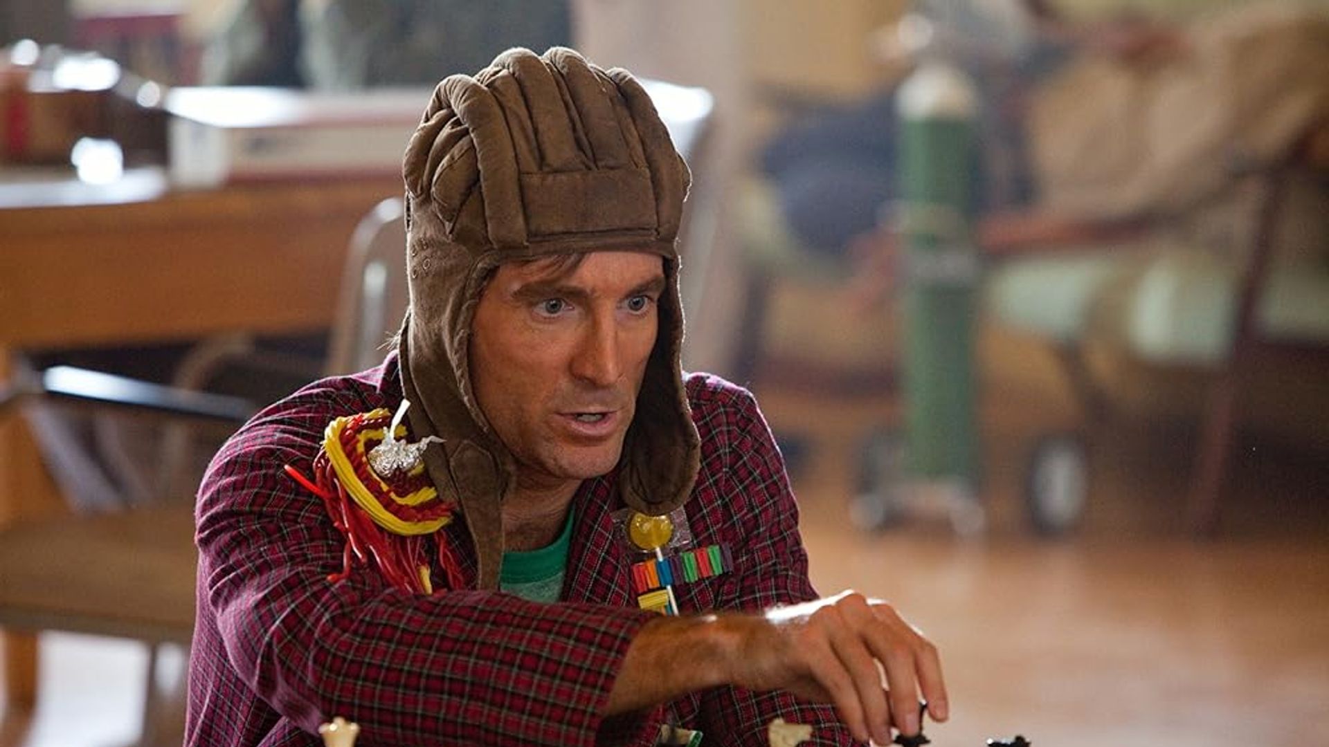 Шарлто Копли в шапке лётчика и красной рубашке играет в шахматы