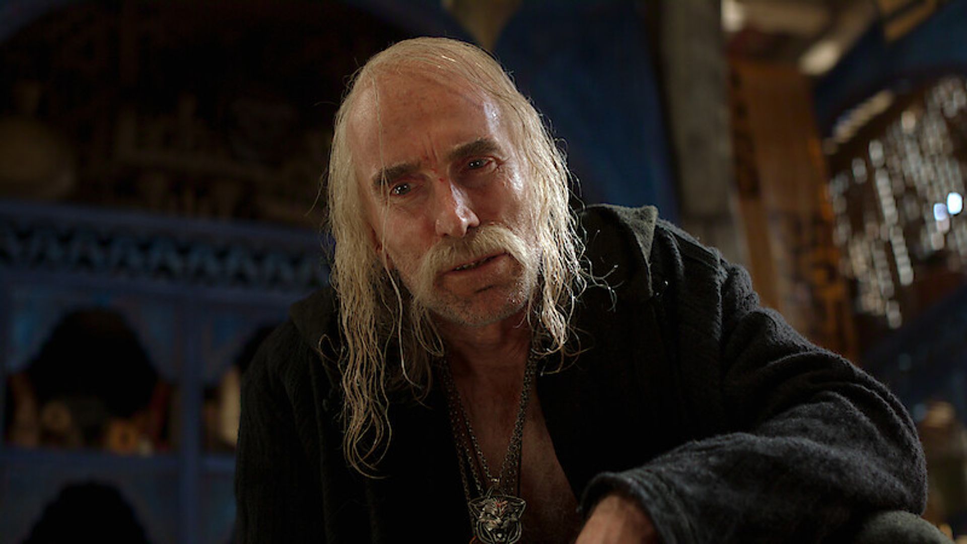 Шарлто Копли с длинными волосами и с медальоном ведьмака, ракурс снизу