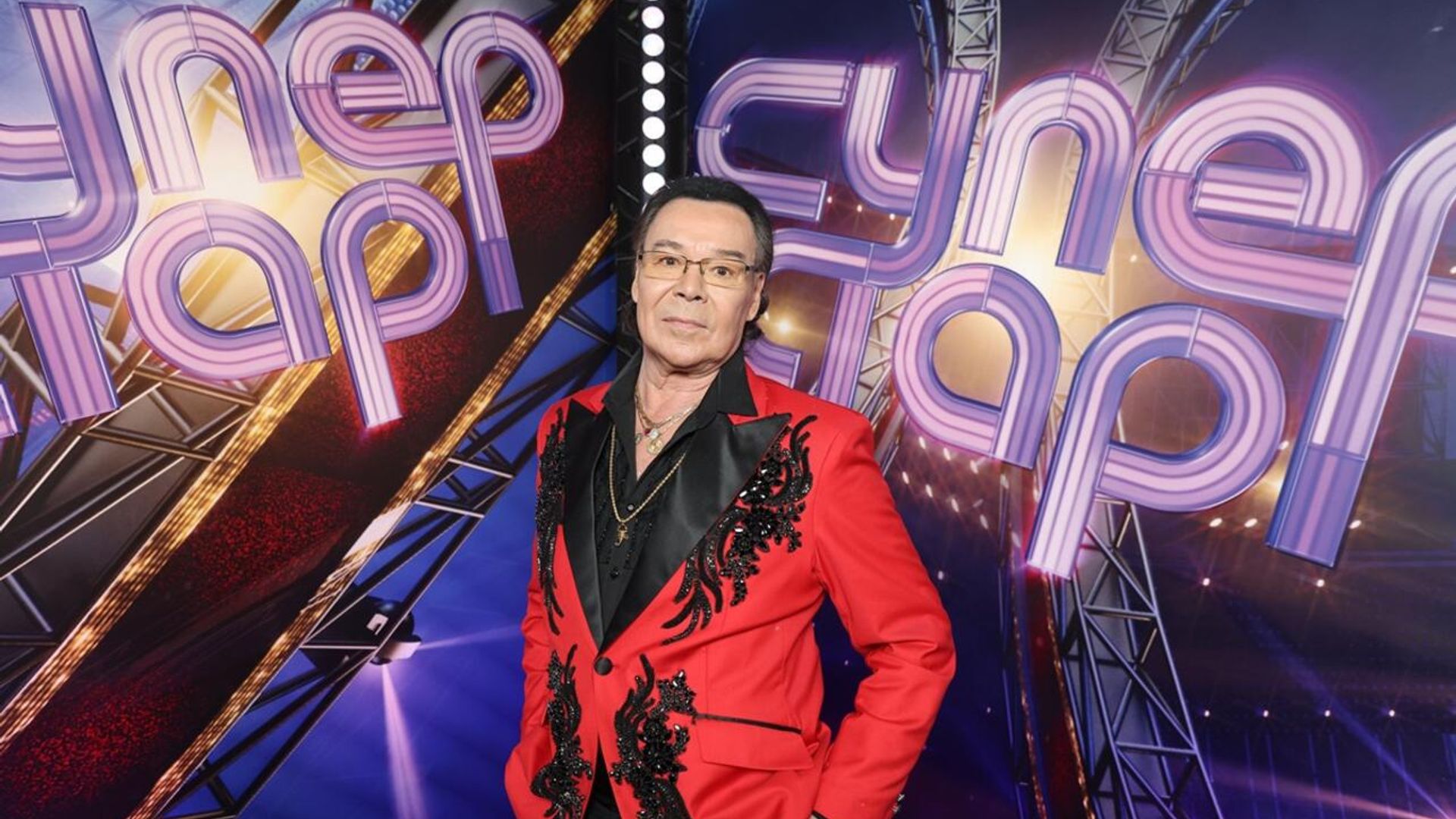 Михаил Муромов в красном сценическом костюме позирует под неоновой надписью "Суперстар"