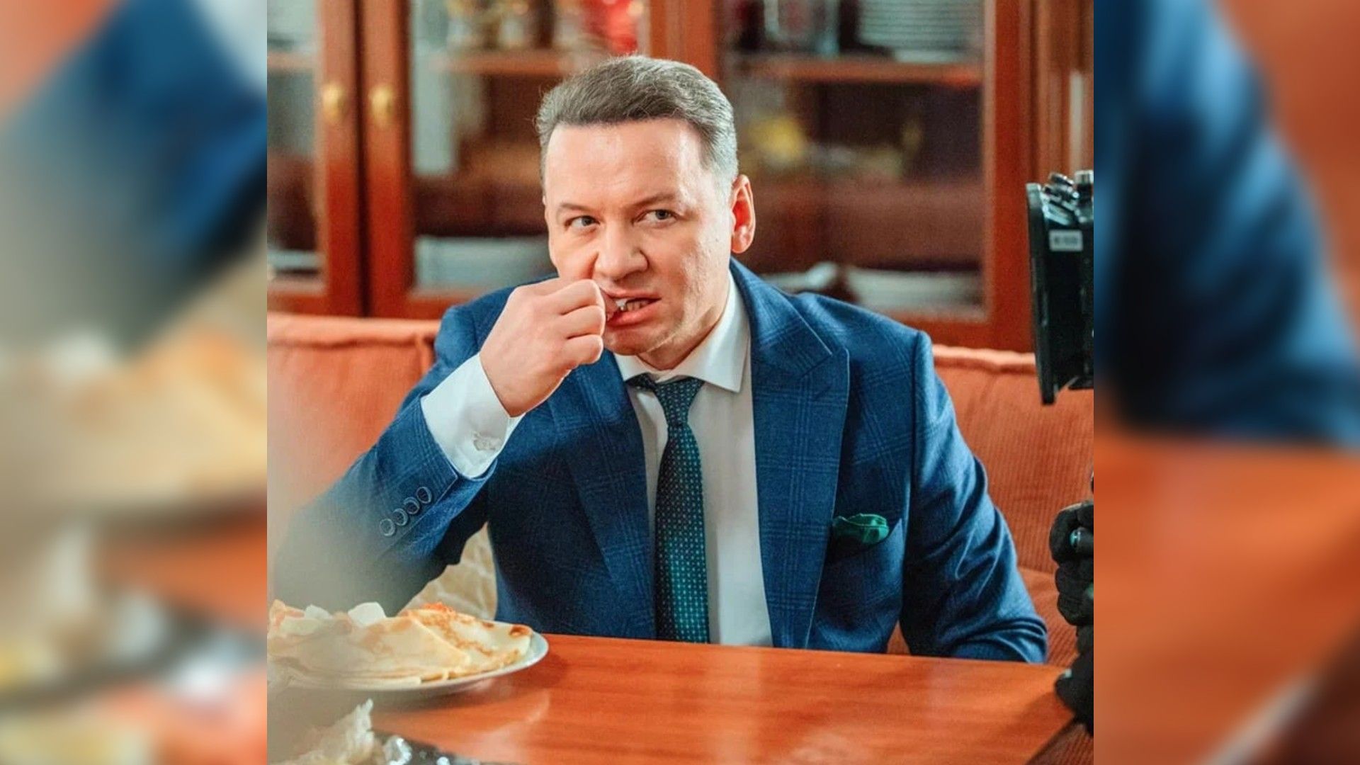 Александр Олешко в костюме сидит за столом и ковыряется в зубах