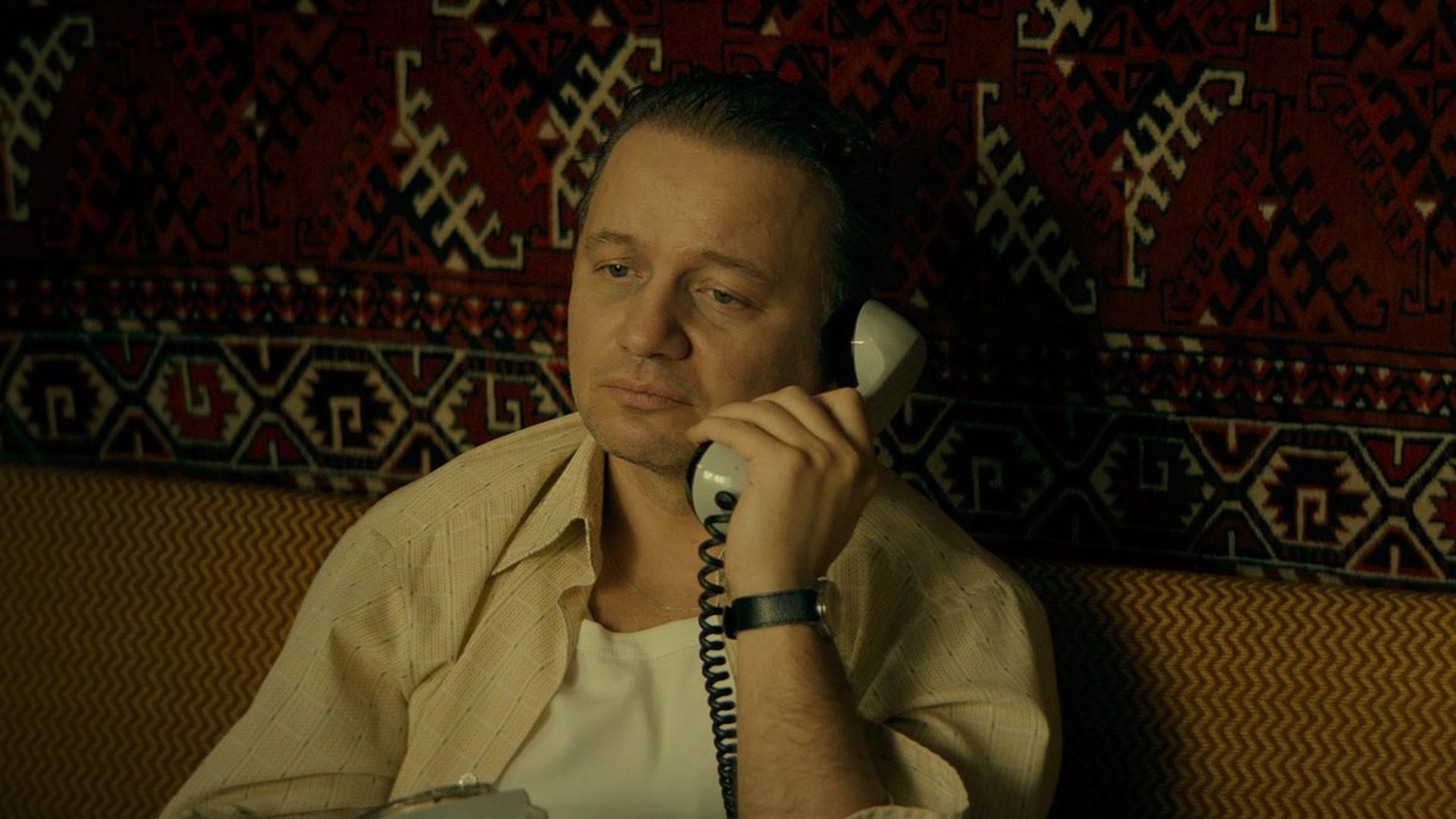 Александр Олешко с грустным выражением лица разговаривает по телефону на фоне советского ковра