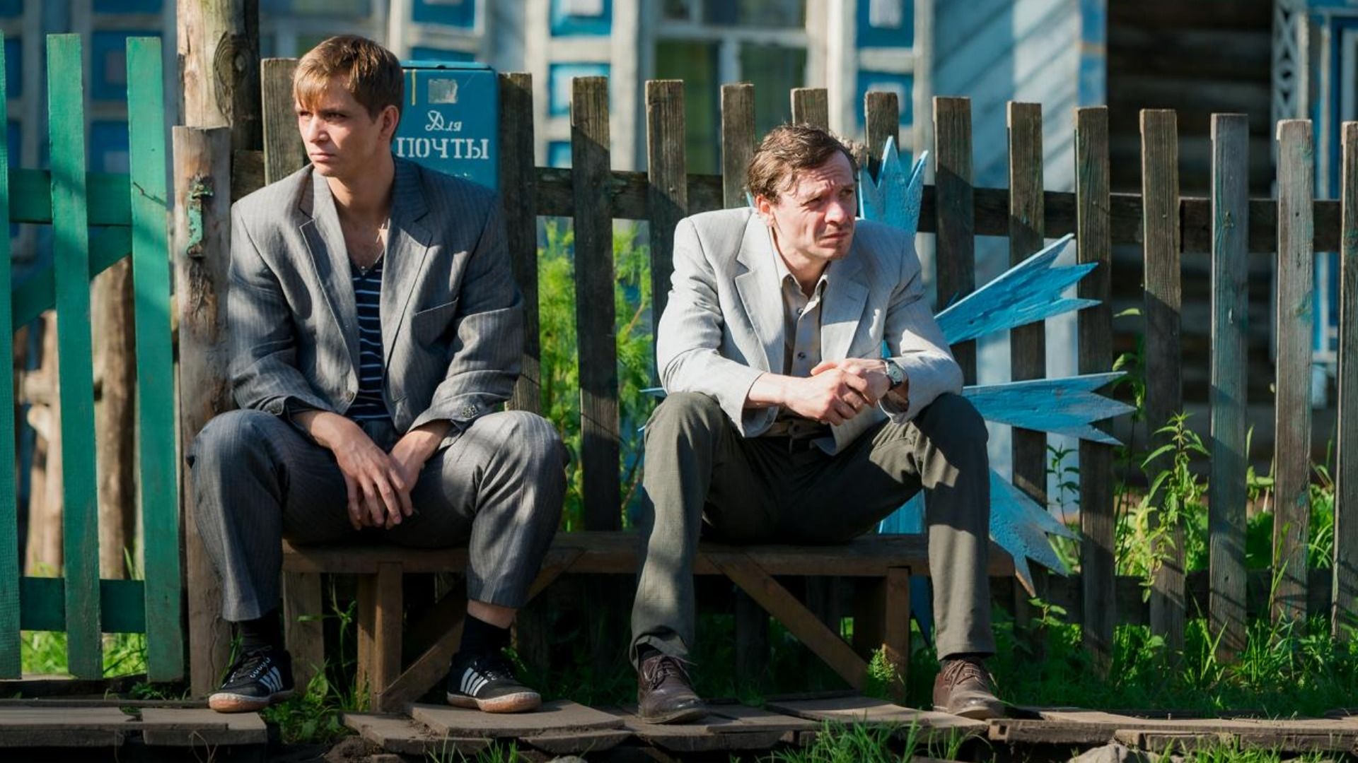 Двое мужчин в серых пиджаках сидят на лавочке у дома