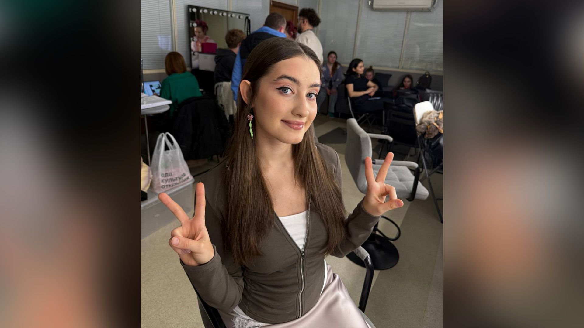 Динара Курбанова сидит на стуле в комнате, полной людей, и показывает пальцами жест V
