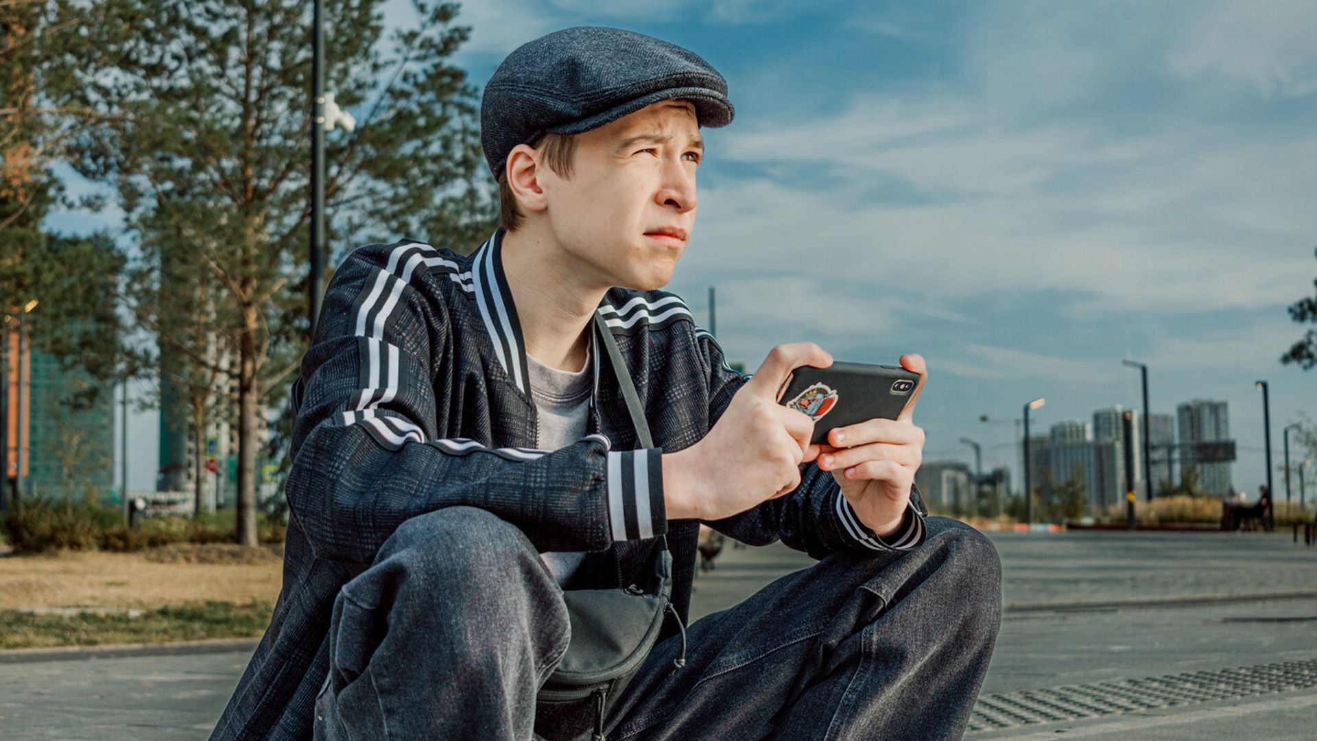 Григорий Столбов в кепке сидит на бордюре, держа в руках смартфон