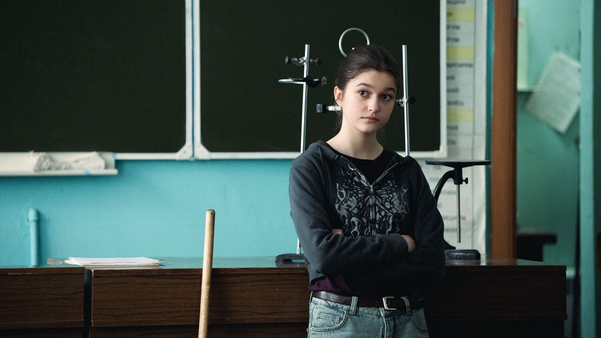 Василиса Коростышевская, скрестив руки на груди, опирается на стол учителя в школьном классе