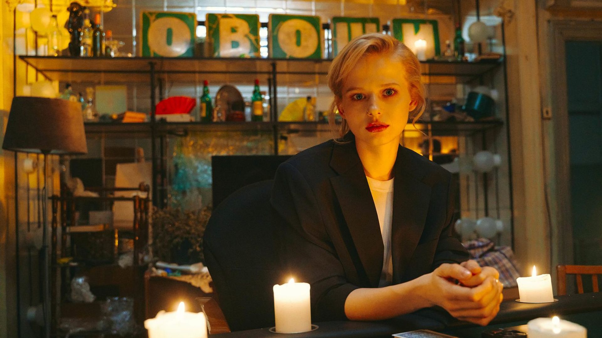 Александра Бортич в пиджаке сидит за прилавком, на заднем плане надпись "овощи"