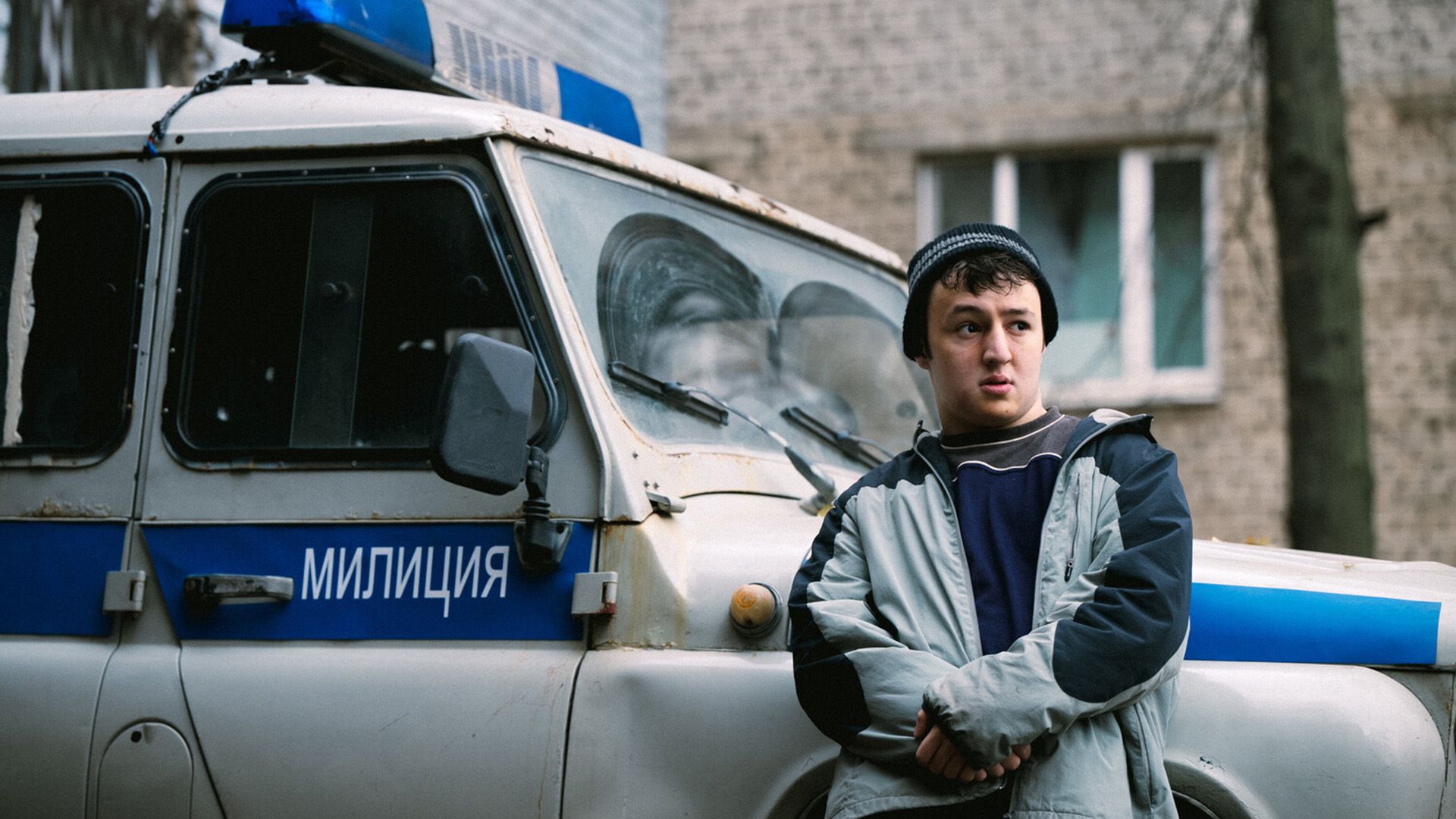 Хетаг Хинчагов в серой куртке облокотился на автомобиль с надписью "Милиция" и недоверчиво смотрит в сторону