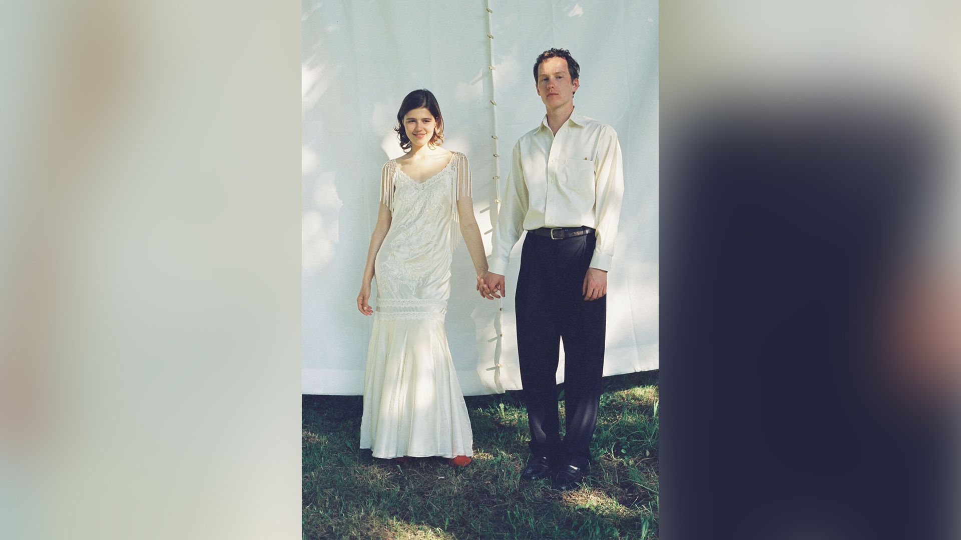 Мария Мацель в свадебном платье и Илларион Маров в брюках и рубашке стоят, держась за руки, на фоне белого занавеса