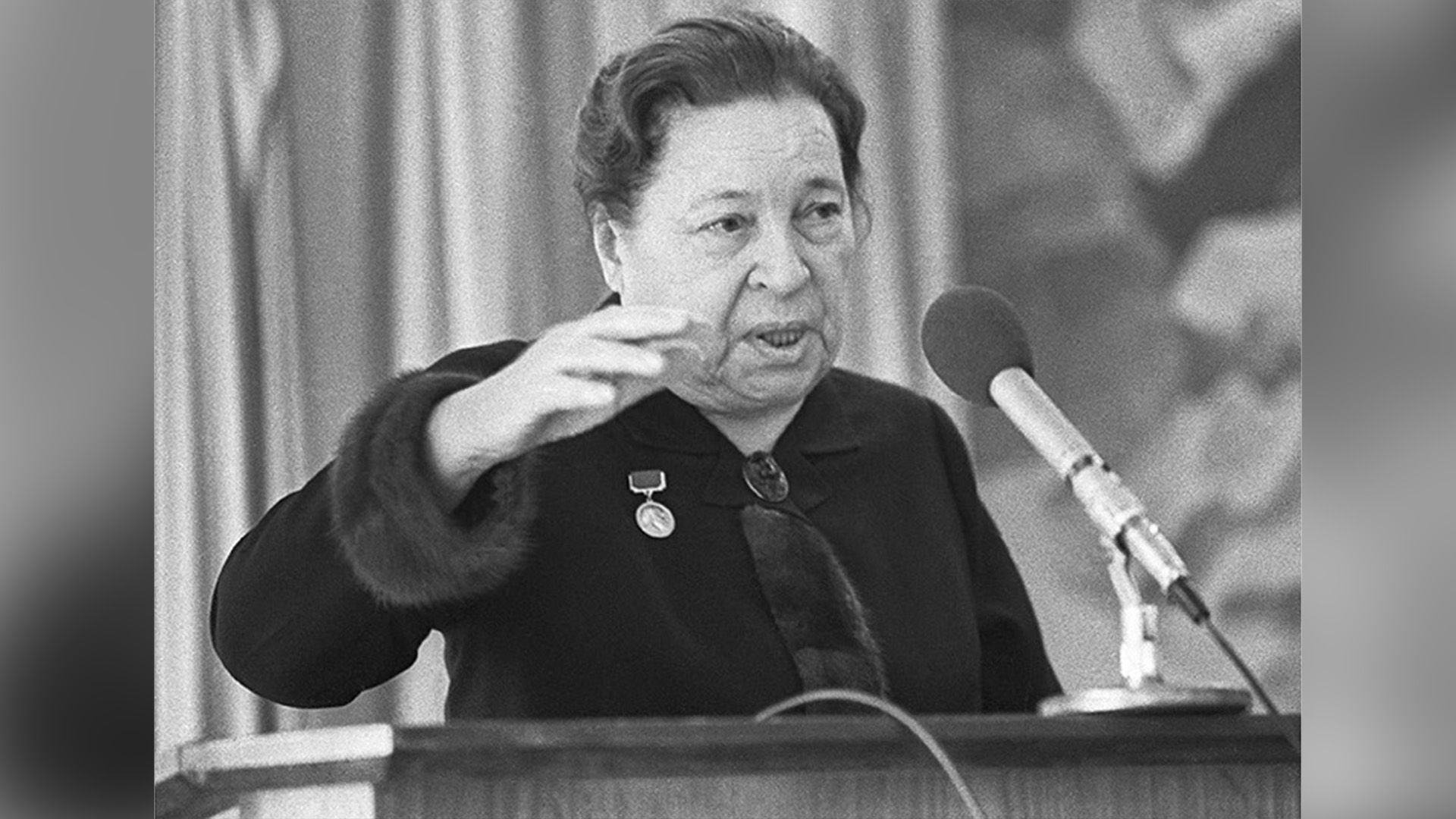 Агния Барто стоит за трибуной и что-то говорит в микрофон, подняв руку