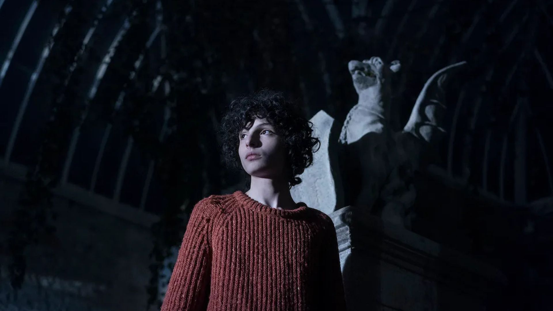 Финн Вулфхард в красном свитере стоит перед статуей гаргульи