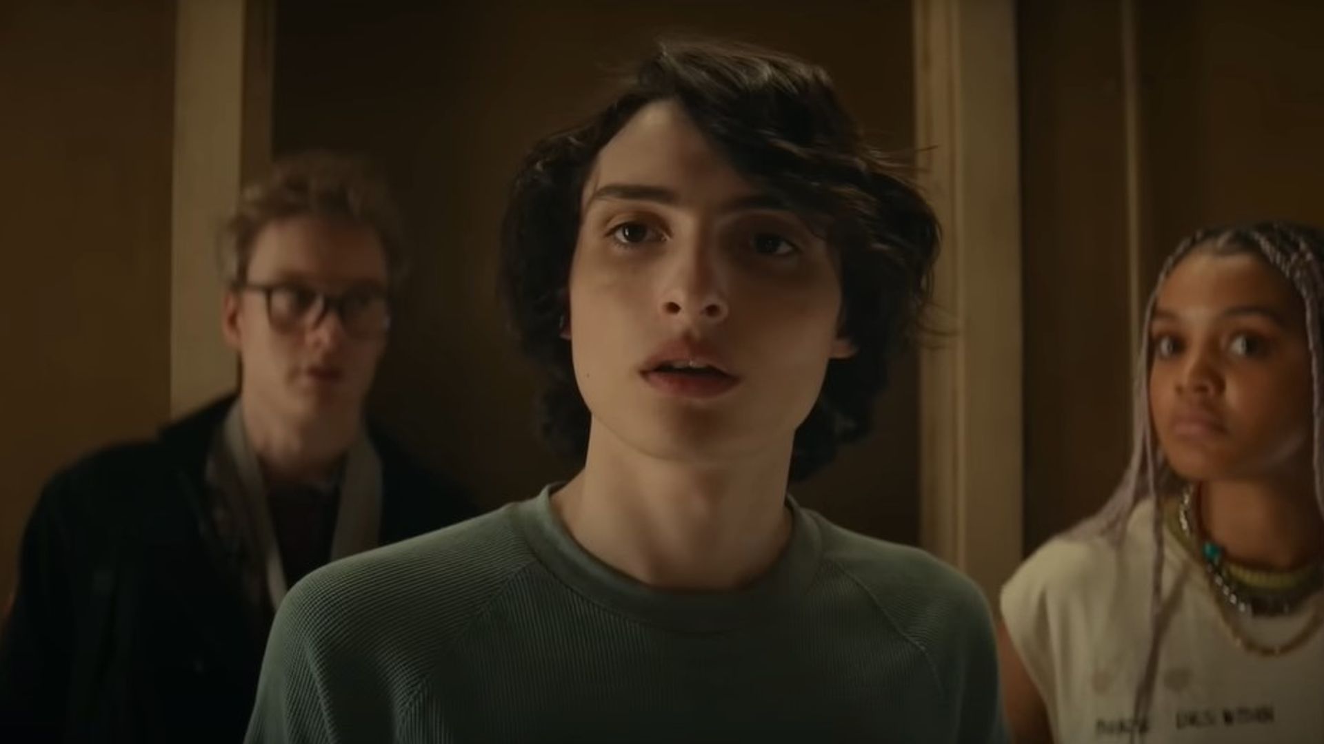 Финн Вулфхард с удивленным выражением лица стоит на фоне двух других людей