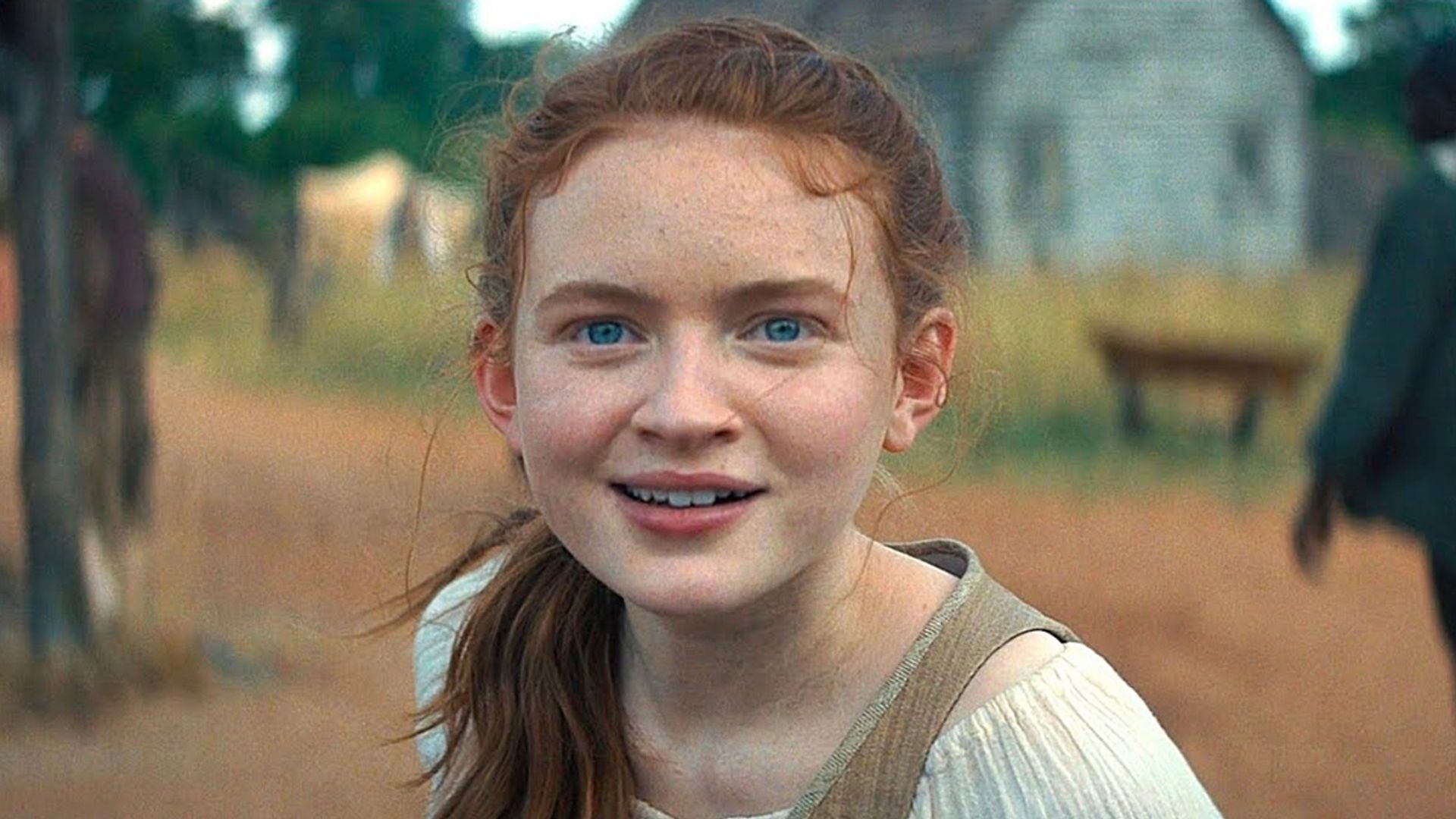 Сэди Синк с хвостиком смотрит прямо в камеру, на заднем фоне деревенский пейзаж