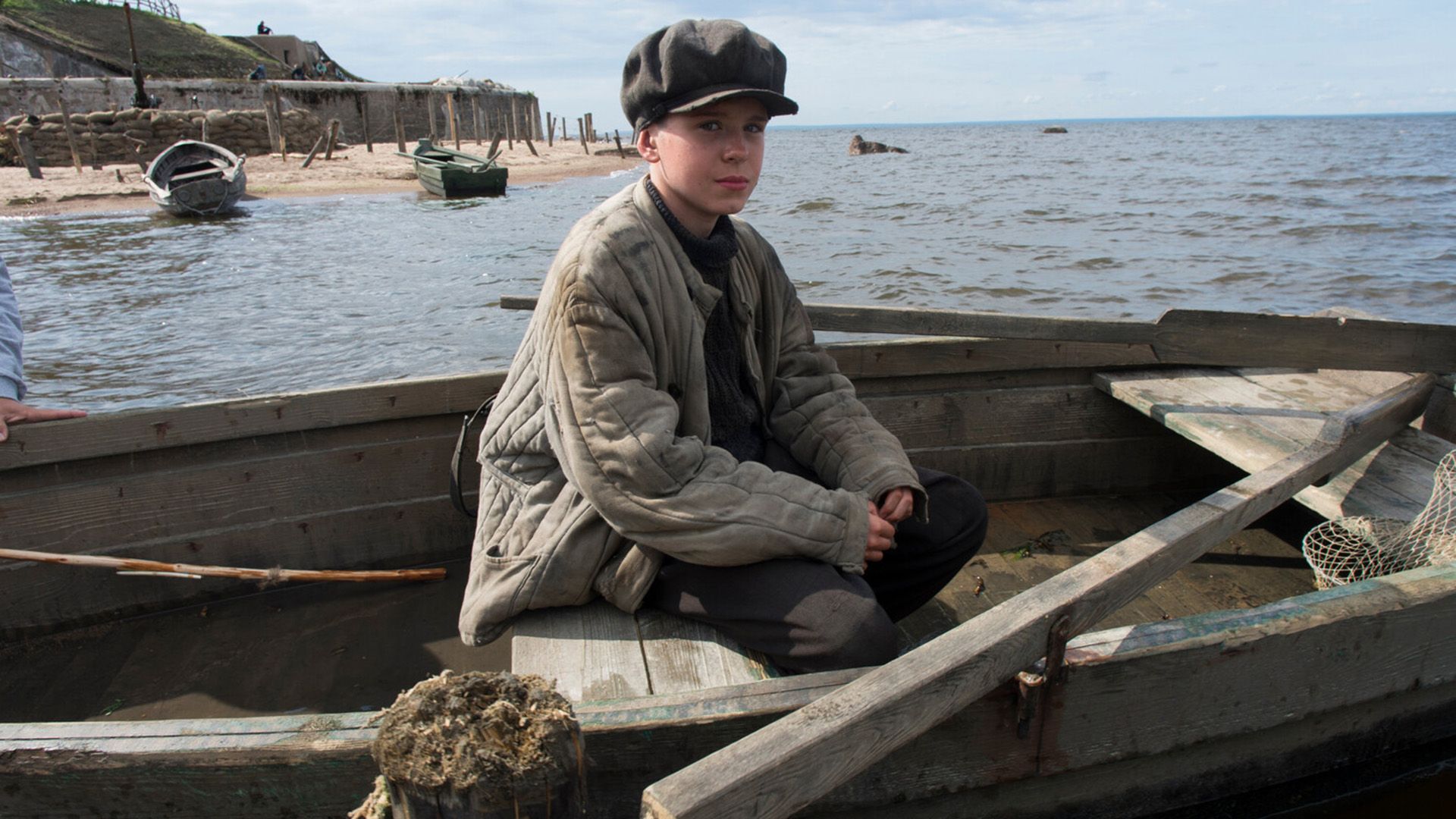 Марк-Малик Мурашкин, одетый в ватник и кепку, сидит в деревянной лодке