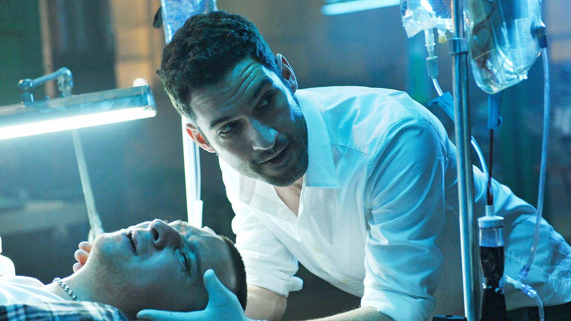 Том Эллис в белой рубашке держит руками голову мужчины, лежащего перед ним, вокруг стоят капельницы