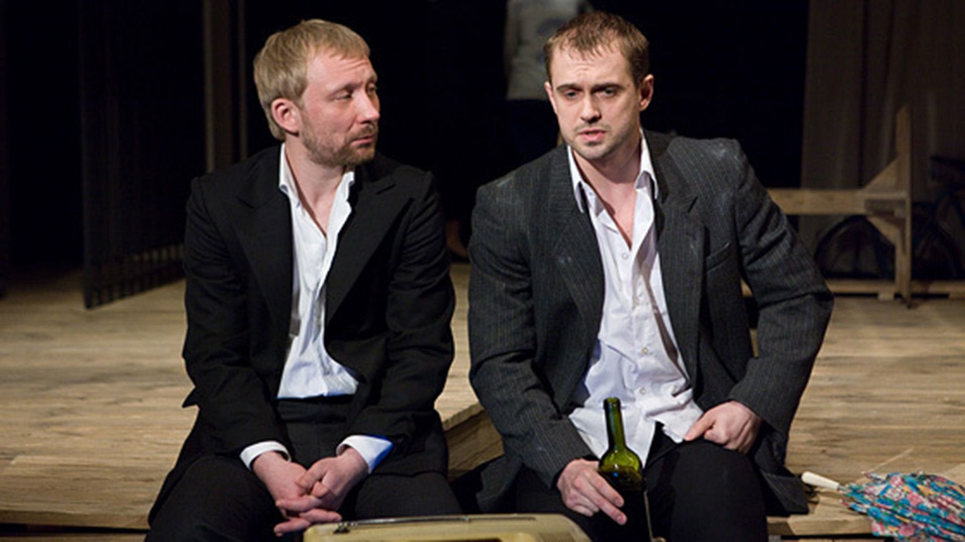 Алексей Комашко в мятом пиджаке сидит на сцене с бутылкой в руке, и что-то рассказывает сидящему рядом мужчине