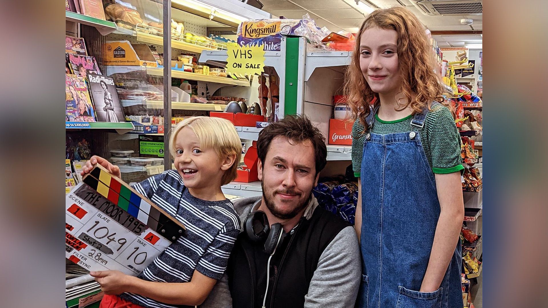 Декстер Сол Анселл с кино-хлопушкой в руках стоит рядом с мужчиной с наушниками на шее и девочкой в джинсовом сарафане