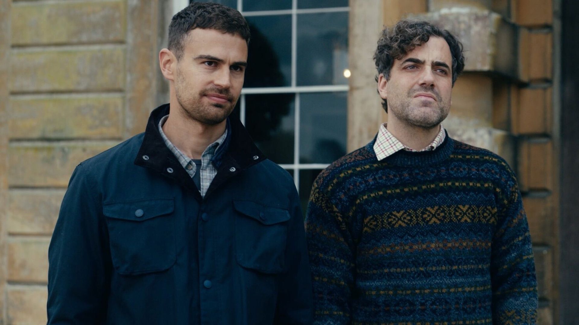 Тео Джеймс в синей куртке и Дэниел Ингс в свитере с орнаментом стоят с хмурыми лицами у кирпичного дома