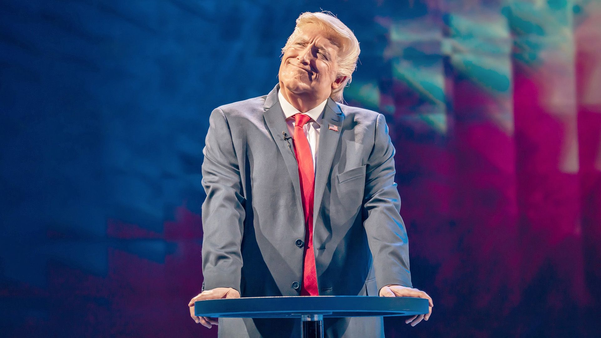 Берти Карвел в образе Дональда Трампа стоит на сцене, оперевшись руками на трибуну