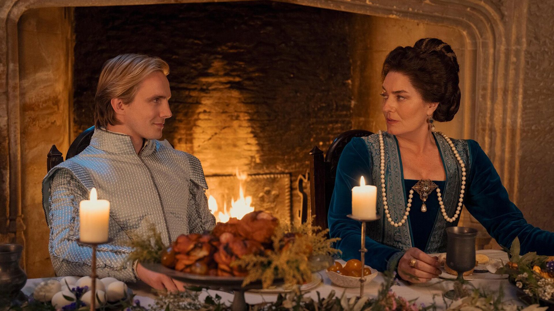 Генри Эштон в голубой костюме середины 16 века сидит за накрытым столом рядом с дамой в синем платье, позади них горит камин