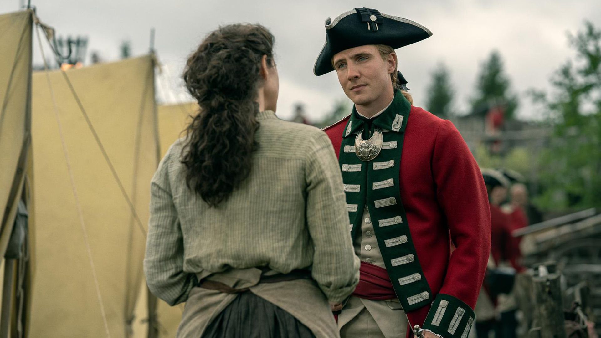 Генри Эштон в красно-зелёном пальто середины 18 века и в треуголке разговаривает с девушкой на фоне военных палаток