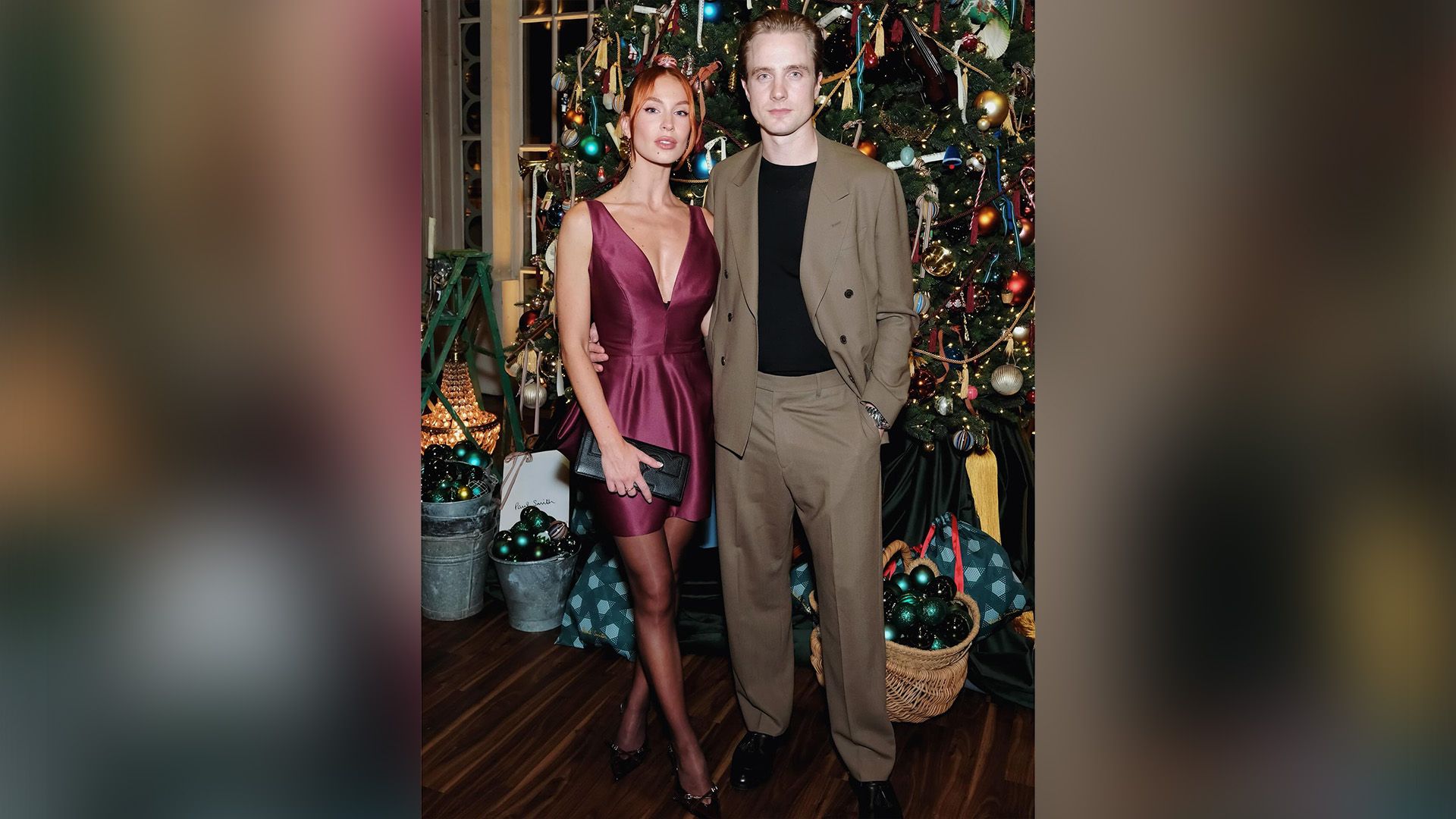 Генри Эштон в светло-коричневом костюме и Эмили-Роуз Бест в бордовом коктейльном платье позируют на фоне рождественской ели