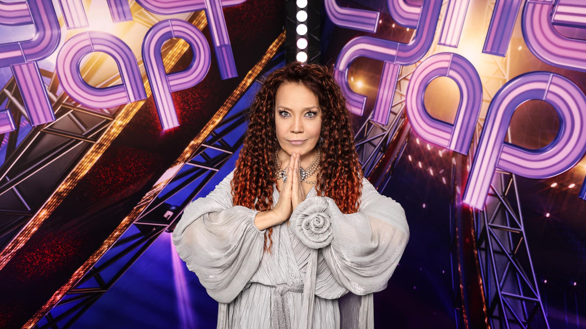 Азиза стоит, сложив руки в молитвенном жесте, на фоне надписи "Суперстар"