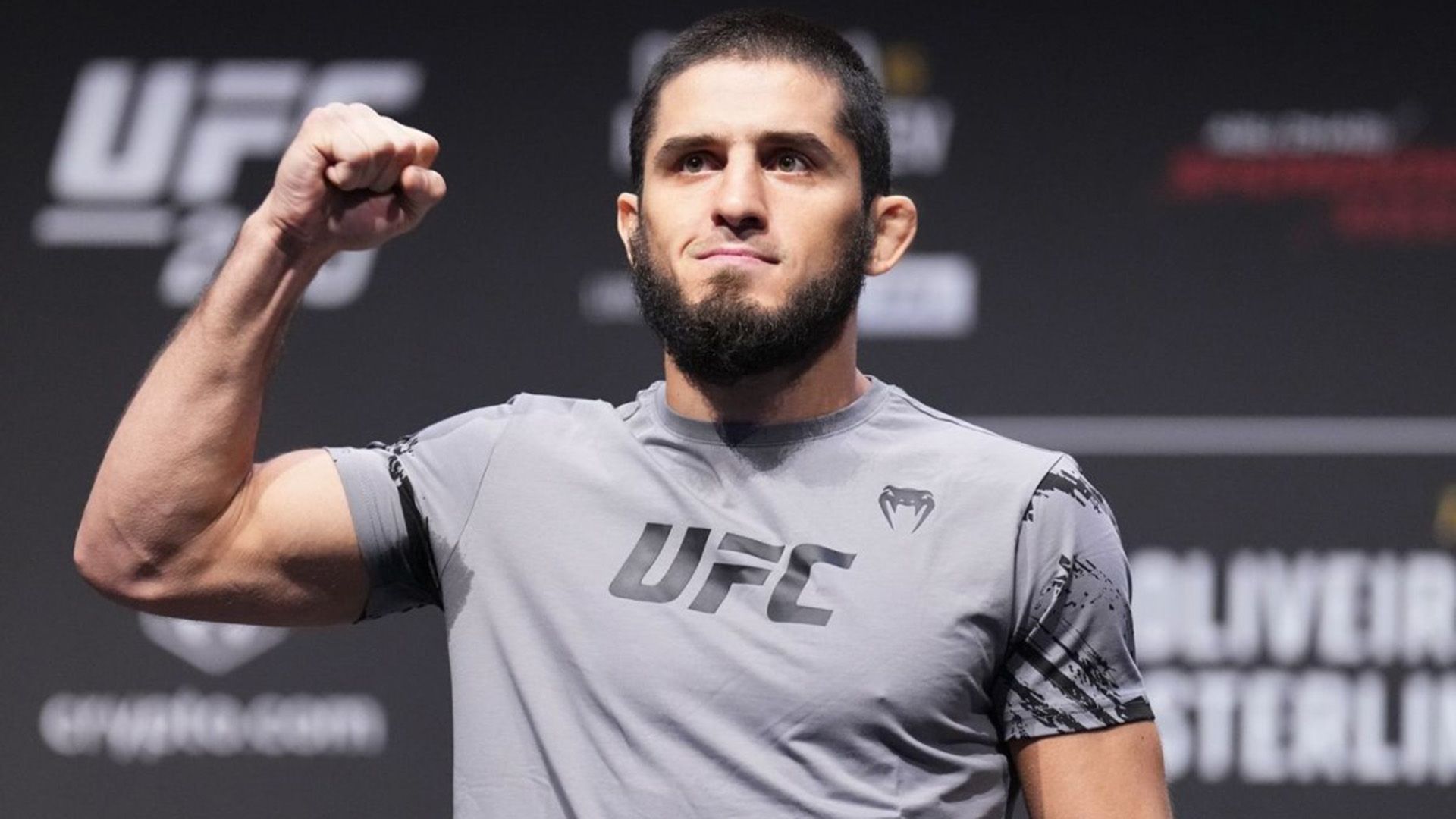 Ислам Махачев в серой футболке стоит с поднятой согнутой правой рукой