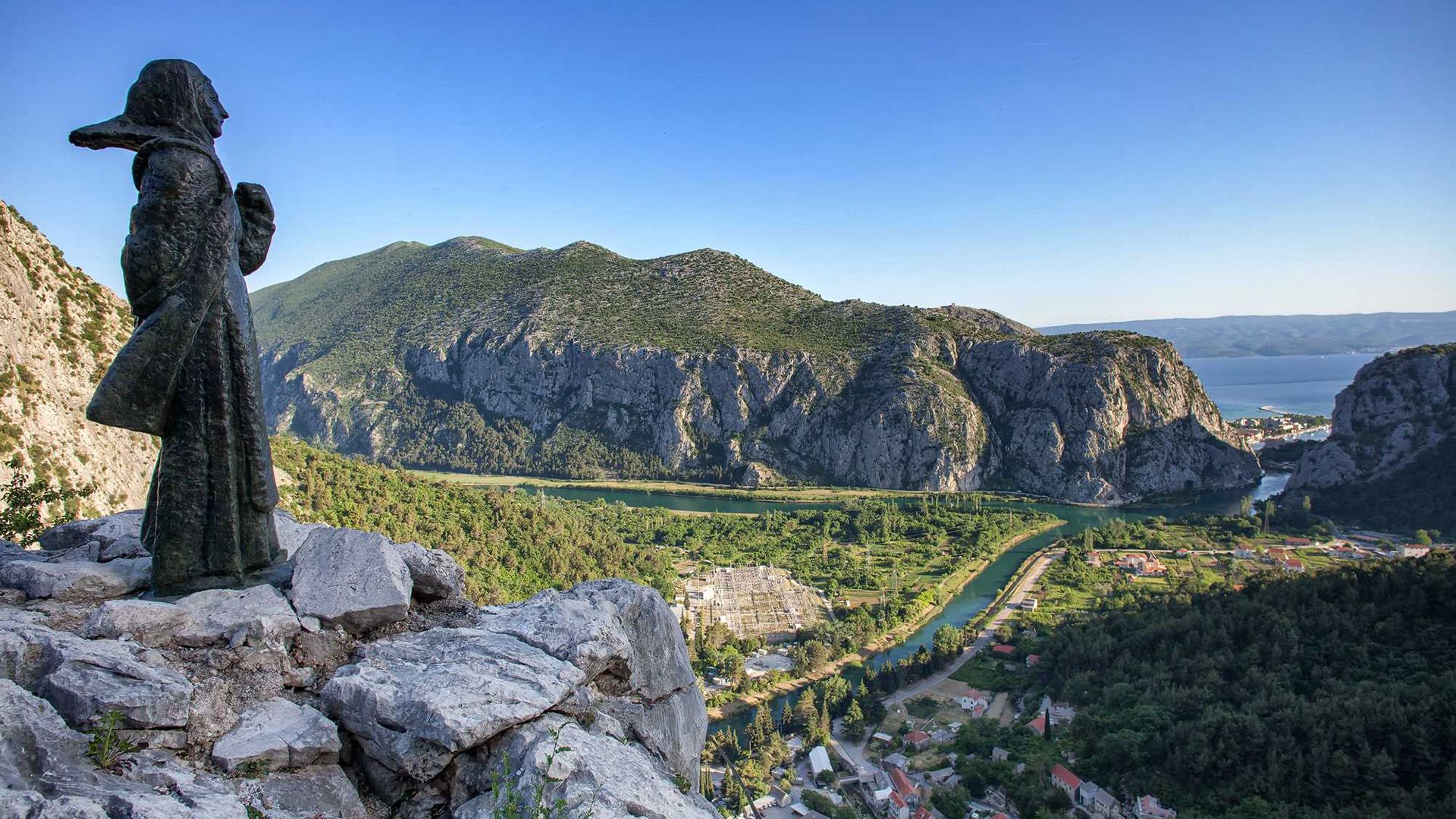 Вид со скалы на маленький город с красными крышами, утопающий в зелени, на скале памятник, изображающий фигуру девушки, смотрящей на город