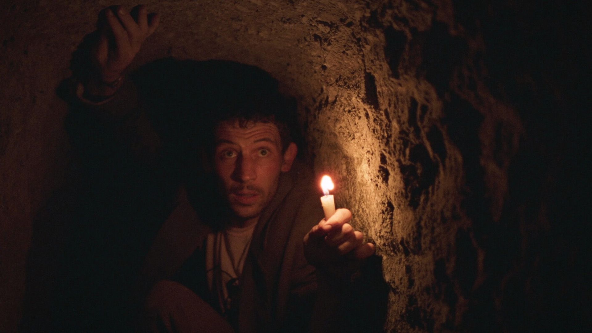 Джош О'Коннор в тесной пещере держит зажжённую зажигалку в вытянутой руке