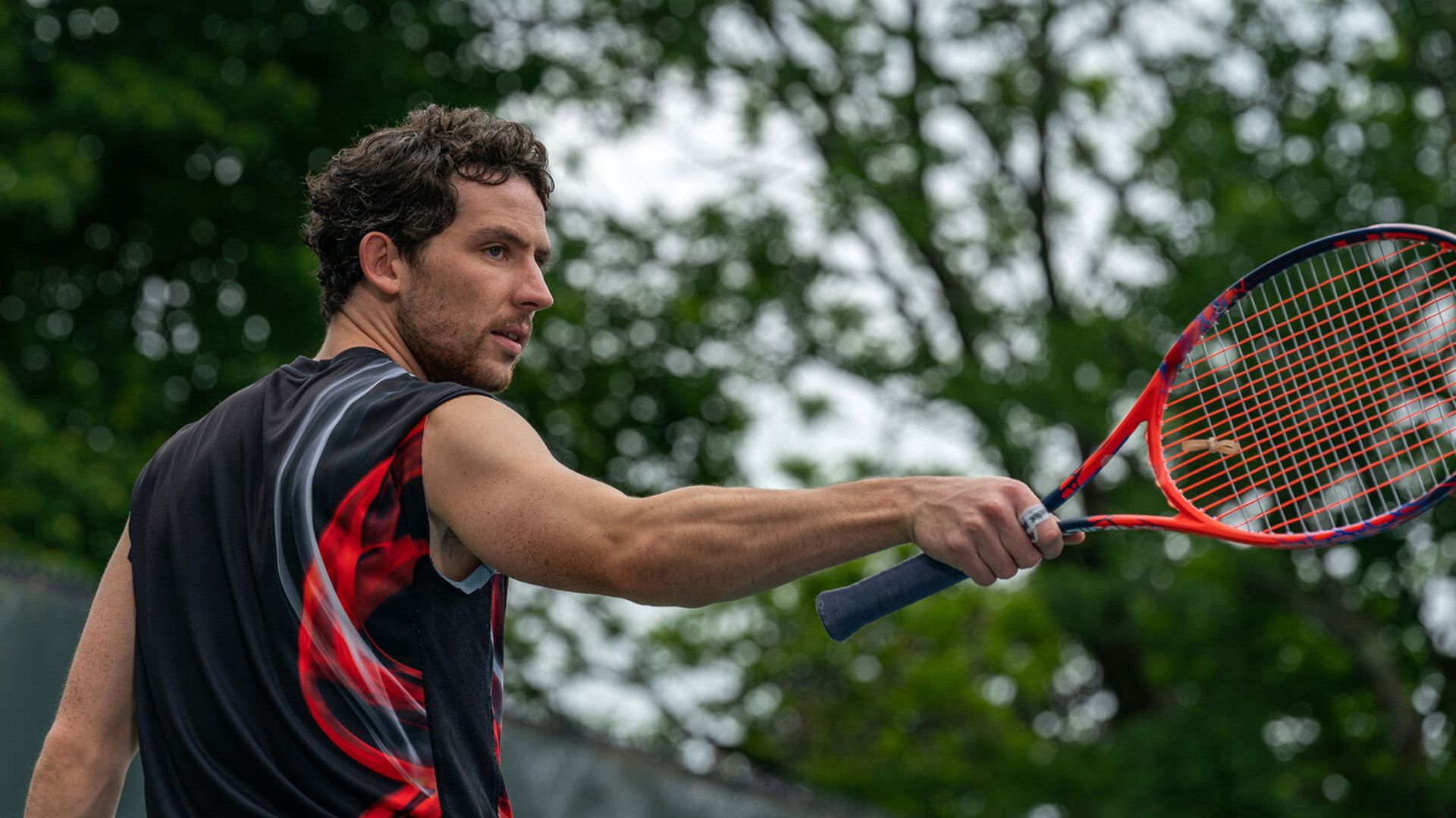 Джош О'Коннор в спортивной чёрно-красной майке и с теннисной ракеткой в руке