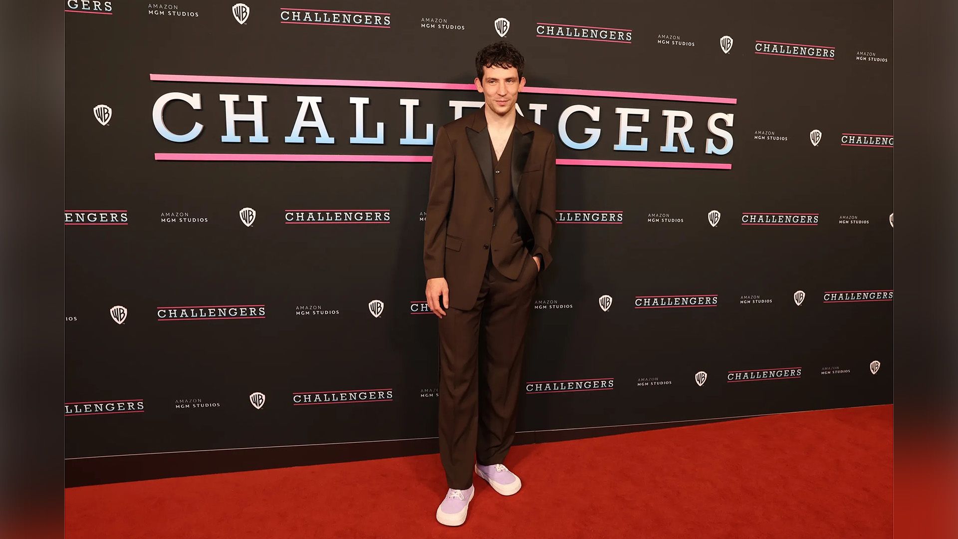 Джош О'Коннор в коричневом классическом костюме и кедах позирует на красной ковровой дорожке